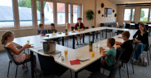 Kinder und Betreuerin sitzen beim Mittagessen an einem U-förmigen Tisch in einem hellen Raum.