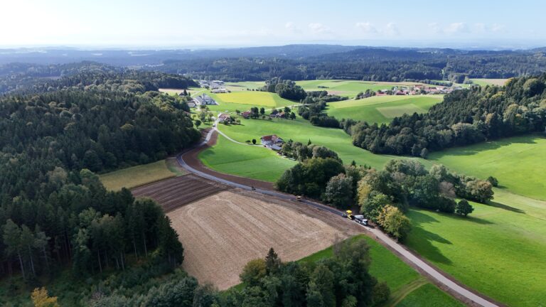 Luftaufnahme der Asphaltierungsarbeiten am Agrarweg Reiting-Kiriau mit Walzen, Asphaltfertiger und Lkw, im Hintergrund Hügellandschaft mit Wäldern und Ortschaft