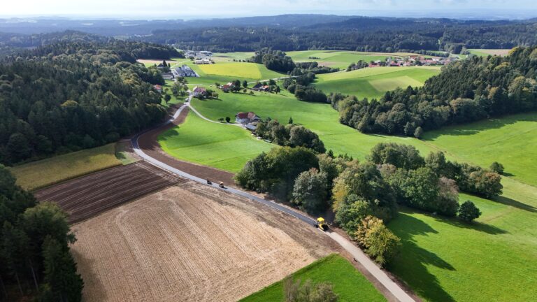 Asphaltfertiger und zwei Walzen auf dem Agrarweg Reiting-Kiriau, Weg entlang eines abgeernteten Feldes und eines Waldrandes